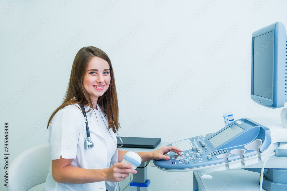 Portrait of young smiling doctor, ultrasound specialist looking at camera and using Ultrasound Scanning Machine for pacient testing. Copy space. Selective focus. - obrazy, fototapety, plakaty 