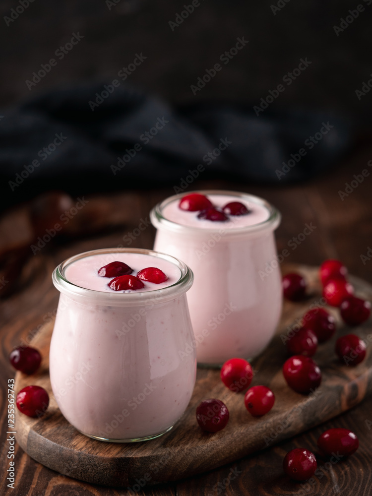 Yogurt with cranberry. Two glass jar with pink yoghurt or milkshake and red cranberries on wooden cutting board over dark background. Copy space for text. Vertical. Yogurt with cranberry. Two glass jar with pink yoghurt or milkshake and red cranberries on wooden cutting board over dark background. Copy space for text. Vertical.