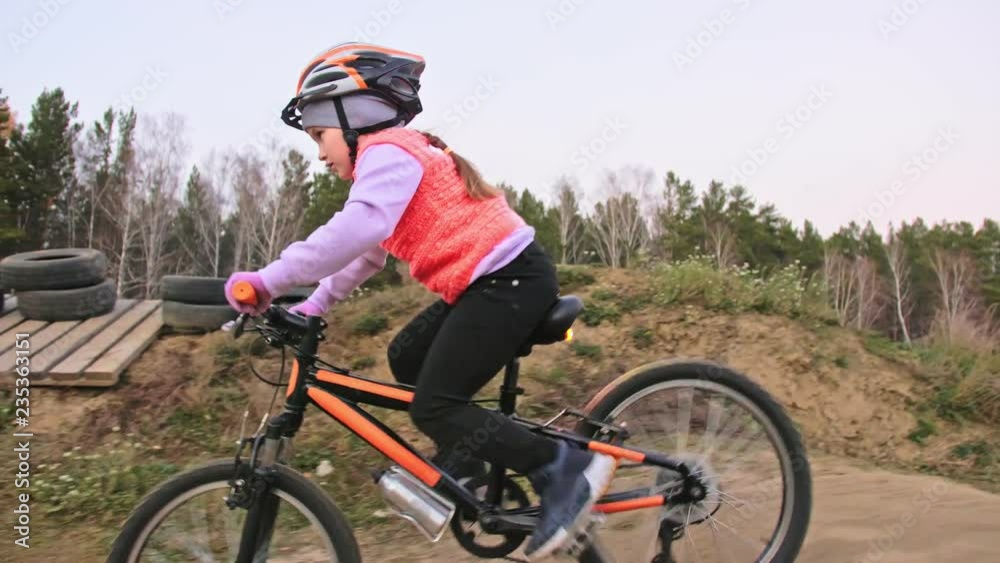 One caucasian children rides bike road track in dirt park. Girl riding black orange cycle in racetrack. Kid goes do bicycle sports. Biker motion ride with backpack and helmet. Mountain bike hardtail. One caucasian children rides bike road track in dirt park. Girl riding black orange cycle in racetrack. Kid goes do bicycle sports. Biker motion ride with backpack and helmet. Mountain bike hardtail.