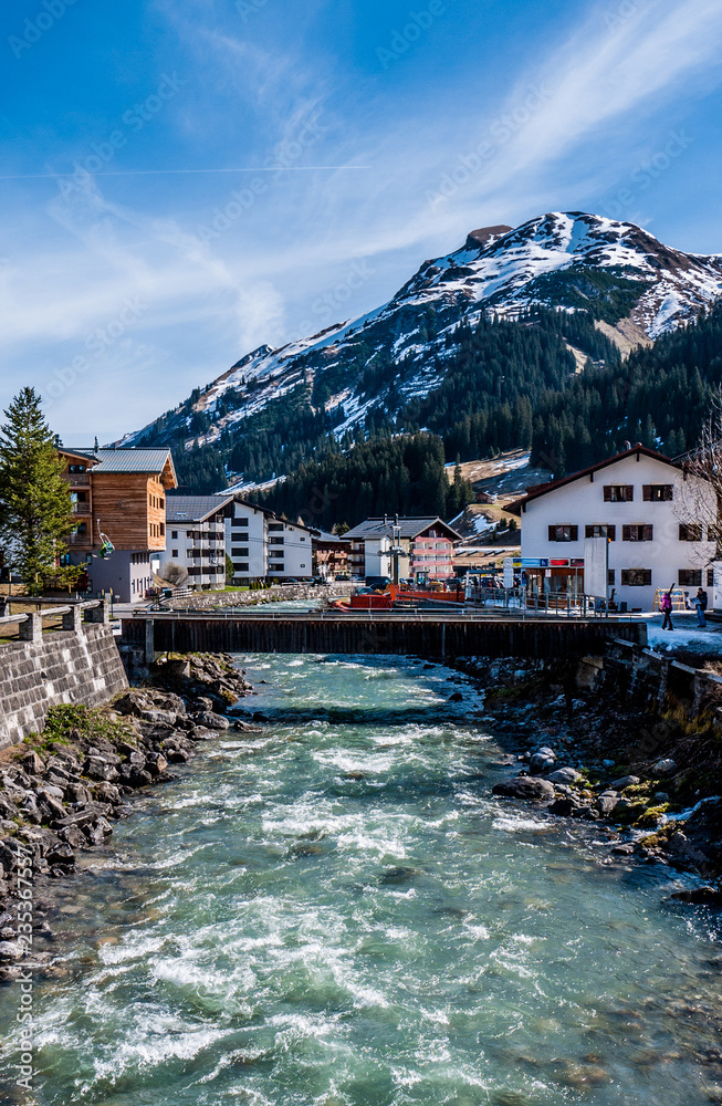 Blue river flowing through Lech in Austria, showing the traditional architecture and tall, snowy mountains.