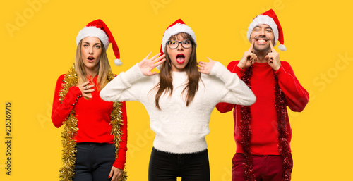 A group of people Blonde woman dressed up for christmas holidays surprised and shocked. Expressive facial emotion on yellow background