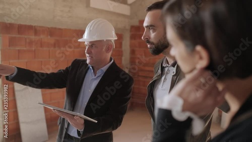 Sales agent talking with clients in new building. Man working as realtor in construction site with customers. Real estate broker showing home to husband and wife. Young couple as buyers of new house