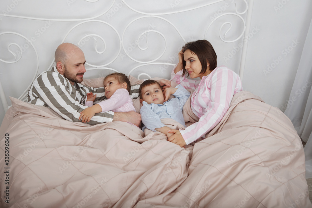 Young pretty woman mother and bald man father with son and daughter in pajamas lie in bed with pillows and pink bedclothes. Young pretty woman mother and bald man father with son and daughter in pajamas lie in bed with pillows and pink bedclothes.