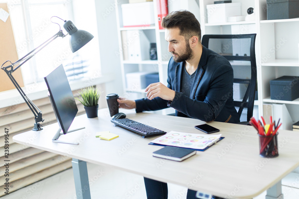 A young man working in the office at the computer and holding a glass of coffee. A young man working in the office at the computer and holding a glass of coffee.