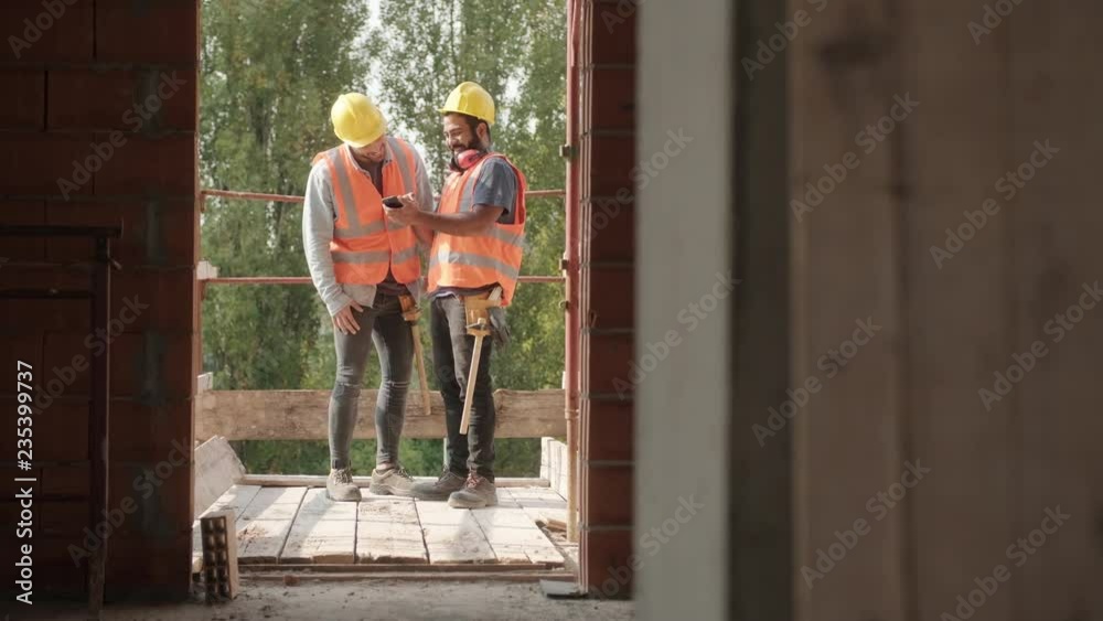 Persons working in construction site. Young men at work in new house inside apartment building. Latino manual workers laughing and using smartphone for internet and social media during break Persons working in construction site. Young men at work in new house inside apartment building. Latino manual workers laughing and using smartphone for internet and social media during break