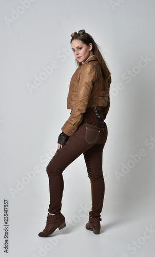 full length portrait of brunette girl wearing brown leather steampunk outfit. standing pose, on grey studio background.