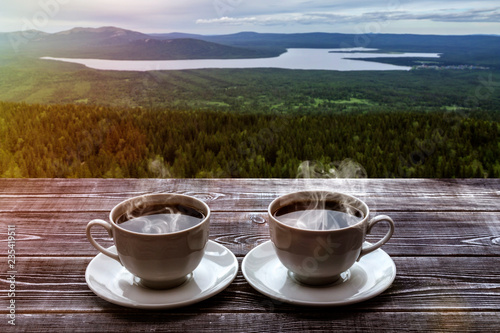 two cups of coffee on a wooden table against a beautiful landscape with a lake and mountains covered with forests. romantic trip.
