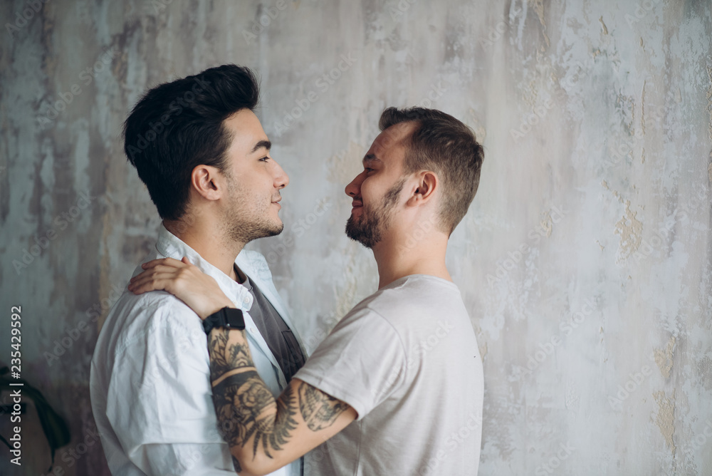 Hot gay couple at the sweet moment of love enjoying closeness and passion kissing isolated over grey lounge rustic background