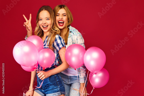 Two smiling beautiful women in checkered shirt clothes.Girls posing on red background.Models with pink balloons.Having fun,ready for celebration birthday or holiday party