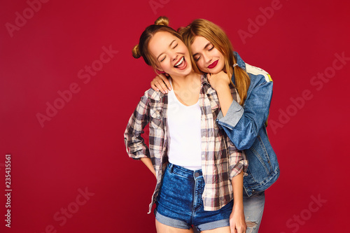 Two young beautiful blond smiling hipster girls posing in trendy summer checkered shirt clothes. Carefree women isolated on red background. Positive models going crazy and hugging