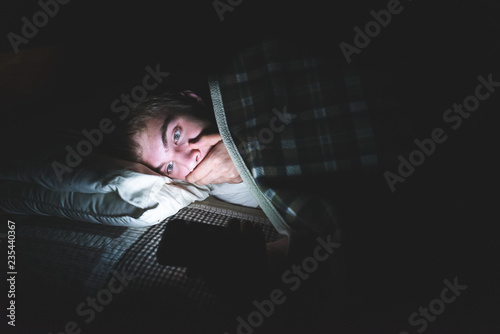 Teenager reading some shocking news on his cell phone as he is lying on his bed in the dark.