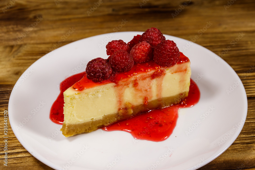 Piece of tasty New York cheesecake with raspberries and raspberry jam in a white plate on wooden table Piece of tasty New York cheesecake with raspberries and raspberry jam in a white plate on wooden table