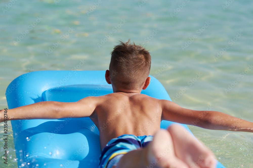 European boy is swimming in the sea on the blue air mattress. He is happy and enjoying his holidays. Back view.