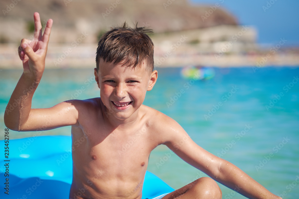 Cute European boy is sitting on a blue air floater in the ocean, smiling and making a 