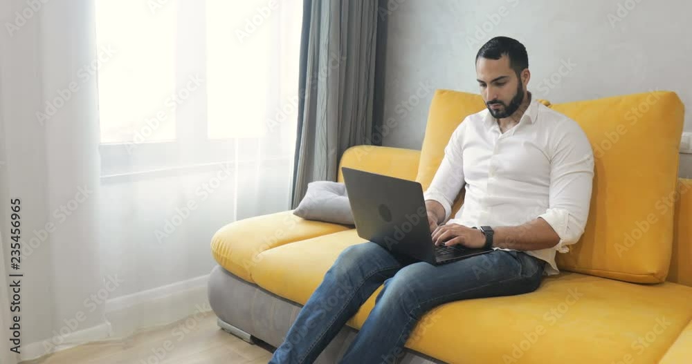 Busy, successful man using laptop as sitting in own living room, wearing pristine white shirt and blue jeans as working from home during lunch break