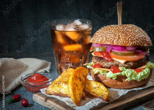 Hamburger with juicy cutlet and vegetables. Spicy sauce and carrots in country style. A glass of coca-cola with ice. On a dark background a blurred background.