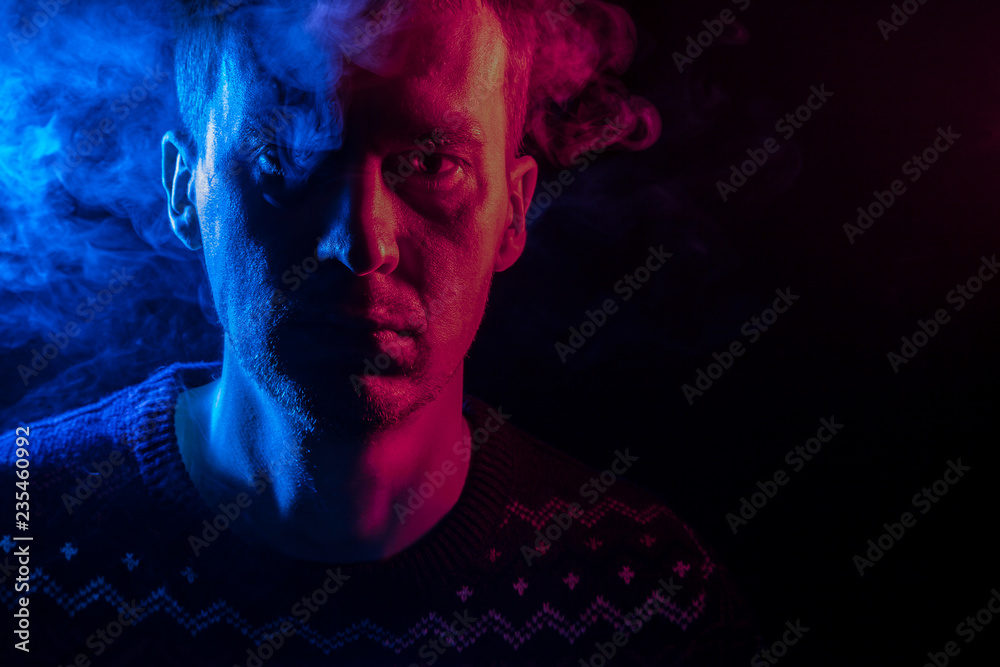 Portrait of a short-haired man with a shadow on a serious face wearing a sweater with colored backlight in blue and pink with smoke around his head. Blurred human mind on black isolated background Portrait of a short-haired man with a shadow on a serious face wearing a sweater with colored backlight in blue and pink with smoke around his head. Blurred human mind on black isolated background