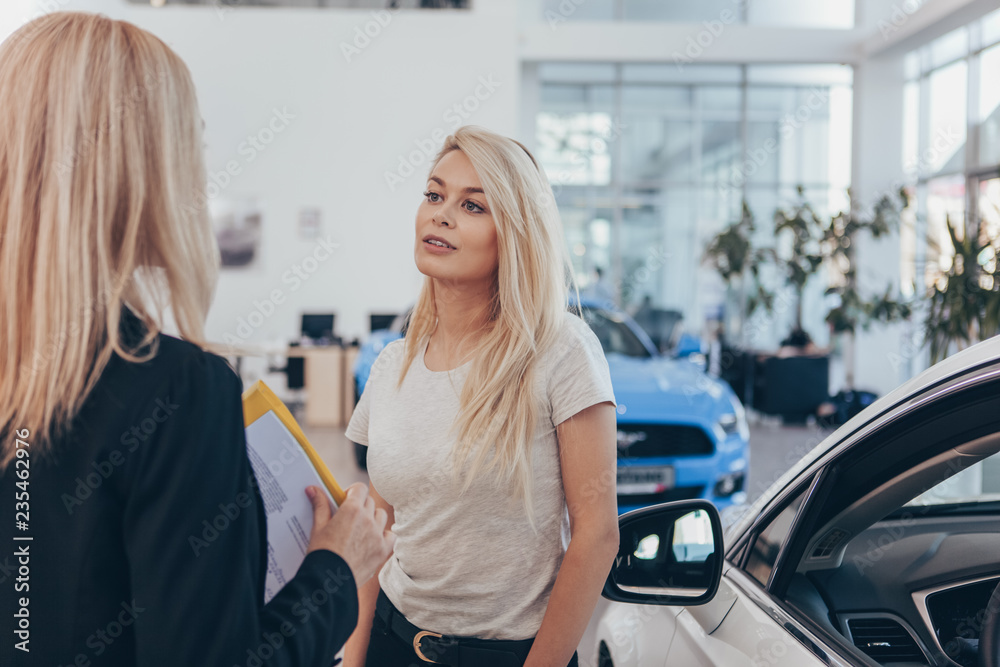 Obraz premium Beautiful woman buying new car at the dealership, talking to the saleswoman, copy space. Professional car dealer helping her client choosing new automobile. Service, promotion, selling concept