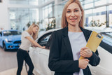 Cheerful mature car saleswoman smiling to the camera, posing with her clipboard in front of new automobile. Female car dealer posing confidently, copy space. Woman choosing new auto on the background