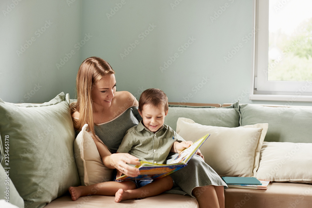 Young mother, read a book to her child, boy in the living room of their home, rays of sun going through the window