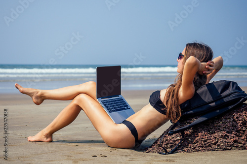 Young woman freelancer in black sexy bikini working on the beach .remote work,freedom and freelancing.successful business girl sitting on the sand in a paradise island working at laptop spf sunscreen