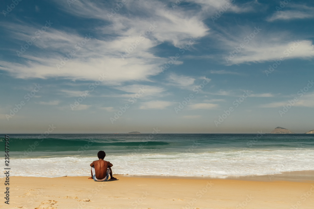 Young African American man sitting alone facing the sea on Copacabana beach in Rio de Janeiro, Brazil