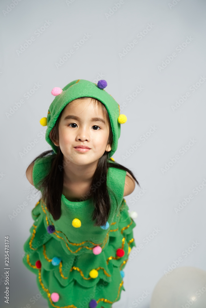 Charming Asian little girl in a christmas tree costume in a studio on white background. She is facing to camera and smile. Happy kid enjoy xmas party. New year holidays eve of Christmas wish concept. Charming Asian little girl in a christmas tree costume in a studio on white background. She is facing to camera and smile. Happy kid enjoy xmas party. New year holidays eve of Christmas wish concept.