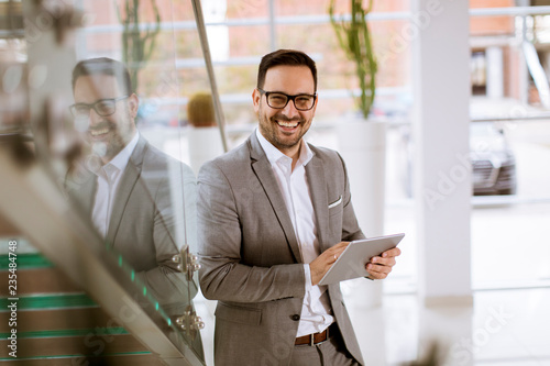 Happy businessman dressed in suit standing  in modern office  and using tablet