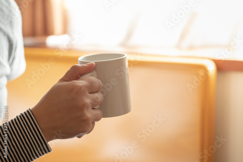 Close up of woman hand holding white mug cup of hot coffee with sunlight in the morning.