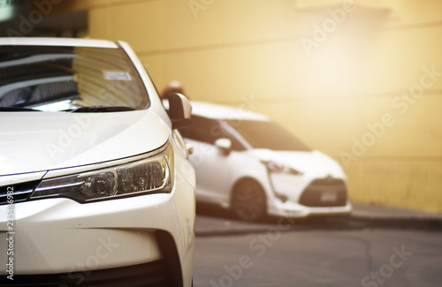 Focusing on the White car headlights on a street corner with sunlight flares and  small white car  In the background  the driver and car. Closeup headlights of car.