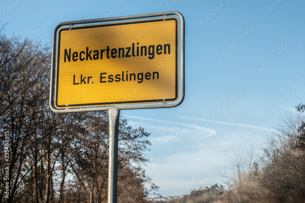 Ortsschild Neckartenzlingen vor blauem Himmel mit Wolken und laublosen Bäumen Ortsschild Neckartenzlingen vor blauem Himmel mit Wolken und laublosen Bäumen
