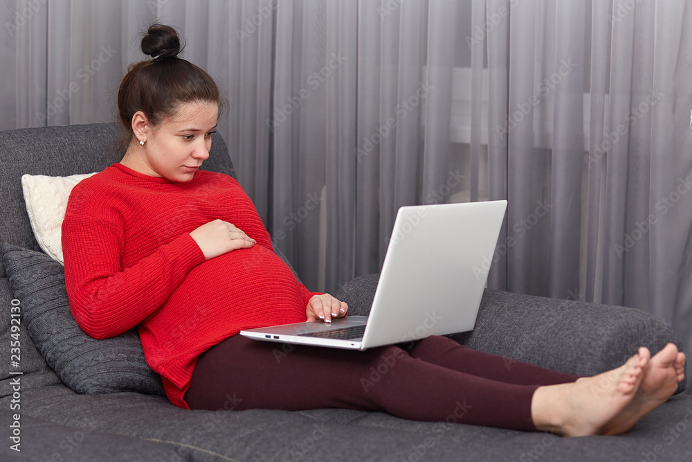 Photo of pregnant woman feels lazy, looks attentively at laptop computer, poses at sofa, watches film online, enjoys free internet connection at home, spends spare time with modern technologies Photo of pregnant woman feels lazy, looks attentively at laptop computer, poses at sofa, watches film online, enjoys free internet connection at home, spends spare time with modern technologies