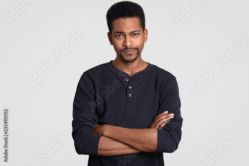 Canvastavla Photo of doubtful serious dark skinned man keeps hands crossed, hears some information, dressed in casual clothes, models against white background