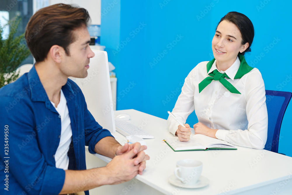 Fototapeta premium Smiling beautiful young lady consultant in green neck scarf sitting at table and making notes while communicating with handsome client
