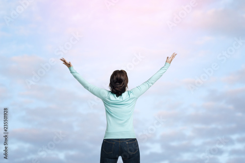 A brunette girl in a mint-colored jumper stands against the background of a delicate blue sky with a lot of small clouds. The young woman raised her hands up to the sky.