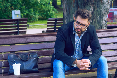 Young man with sad facial expression sitting on a bench in the park. Office worker lost his job. Middle aged man despair economic crisis.