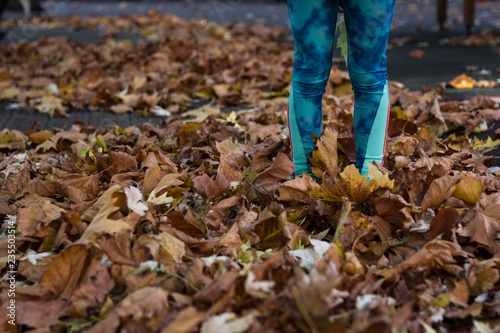 Brown leaves on ground covering feet of little girl in fall season. Child in green leggings standing outdoors on autumn day