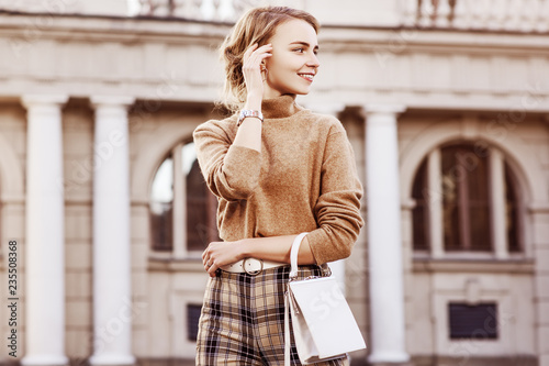 Outdoor fashion portrait of young happy smiling lady wearing trendy beige turtleneck, high-waisted checkered trousers, wrist watch, belt, carrying small white bag, posing in street. Copy, empty space