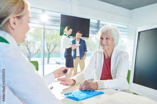 Smiling attractive gray-haired senior lady in white cardigan sitting at table and receiving key from manager while buying flat with mortgage in bank