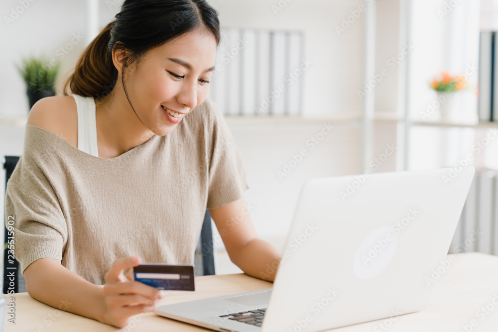 Beautiful Asian woman using computer or laptop buying online shopping by credit card while wear sweater sitting on desk in living room at home. Lifestyle woman at home concept. Beautiful Asian woman using computer or laptop buying online shopping by credit card while wear sweater sitting on desk in living room at home. Lifestyle woman at home concept.