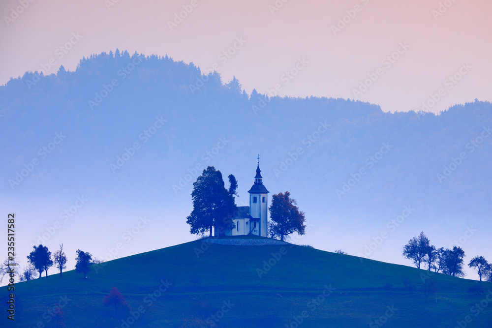 Landscape in Slovenia, nature in Europe.  Foggy Triglav Alps with forest, travel in Slovenia. Beautiful sunrise with blue sky, green nature. Saint Thomas Church, Sveti Tomaz nad Praprotnim.