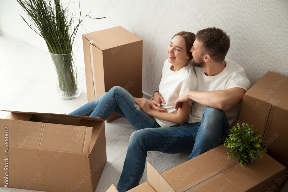 Young laughing couple sitting on floor, embracing in new apartment with packaged belongings in cardboard boxes, family just arrived in new own house, man and woman beginning live together, having fun Young laughing couple sitting on floor, embracing in new apartment with packaged belongings in cardboard boxes, family just arrived in new own house, man and woman beginning live together, having fun