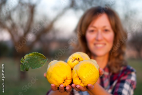 Blonde woman with three quinces in her hand in the foreground, yellow and big, in a field with trees, dressed in a red and white plaid shirt.