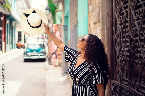 Young beautiful black woman with a typical cuban hat standing in the old streets of Havana Cuba in front of a classic car holding the hat up high. © Saga_bear