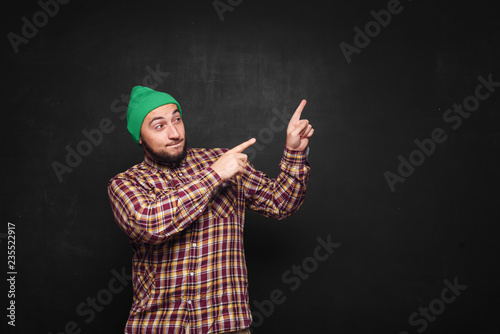 Young european man with beard in green knitted hat thinks, looks surprised and puzzled. Showing fingers upwards and right side. Black background, blank copy space for text or advertisement