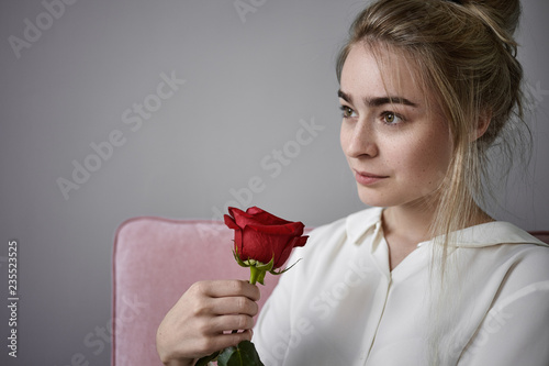 Romance, love and natural beauty concept. Close up cropped view of beautiful romantic young female with fair hair wearing white blouse sitting isolated, smelling red rose on Valentine's Day