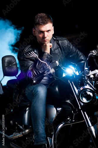 Cute biker in leather jacket sits on a motorcycle in blue smoke 
