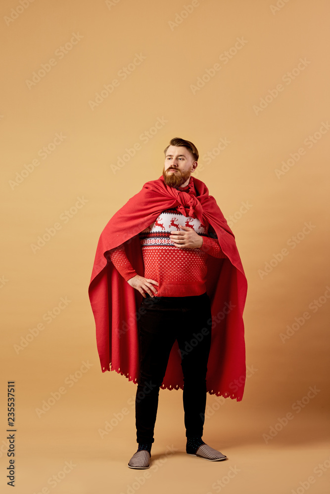 Red-haired man with beard dressed in a red and white sweater with deer and red cape and sleepers stands on a beige background