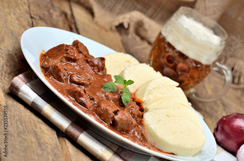 Pork goulash meat with dumplings on white plate, cutlery, cold beer, garlic, onion, pepper, tablecloth in the background - typical Czech food