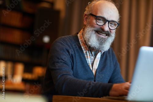 Happy bearded senior man in eyeglasses typing on laptop while browsing in the net